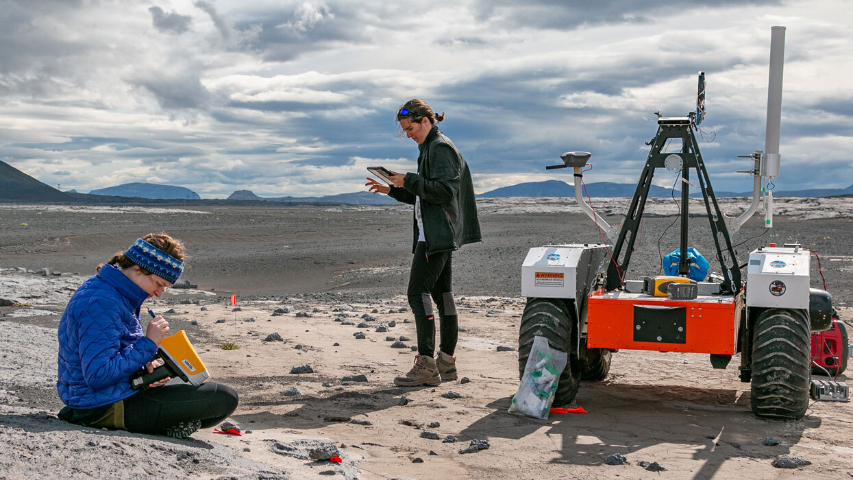 Two geology students conduct research on rocks using technology and a rover.